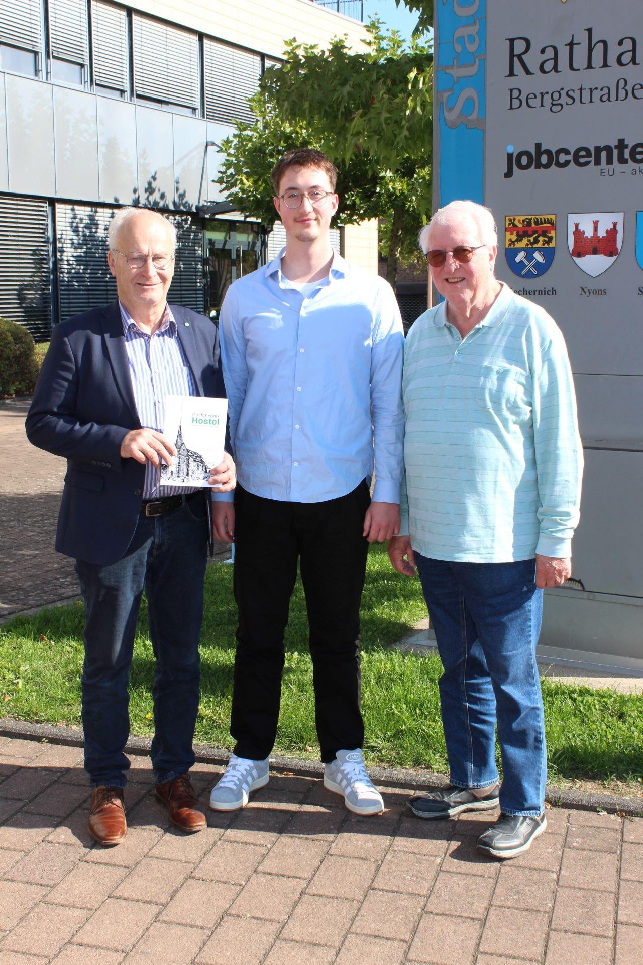 Das erste Exemplar für den ersten Bürger: Noch druckfrisch überreichte Autor Lucas Hamacher (Mitte) die nagelneue Dorfchronik zusammen mit Großvater Dieter Schröder (rechts) an Bürgermeister Hans-Peter Schick, der den 23-Jährigen zu den Recherchen über sein Heimatdorf Hostel ermutigt hatte. (c) Foto: Kerstin Rottland/pp/Agentur ProfiPress