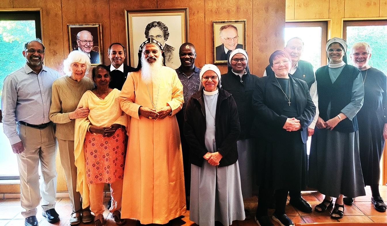 Bischof Dr. Samuel Mar Irenios inmitten der Kommunität der Communio in Christo im Mechernicher Refektorium vor den Bildern der Gründerin Mutter Marie Therese, dem ersten Spiritual Hermann Walch und dem ersten Generalsuperior Karl-Heinz Haus.