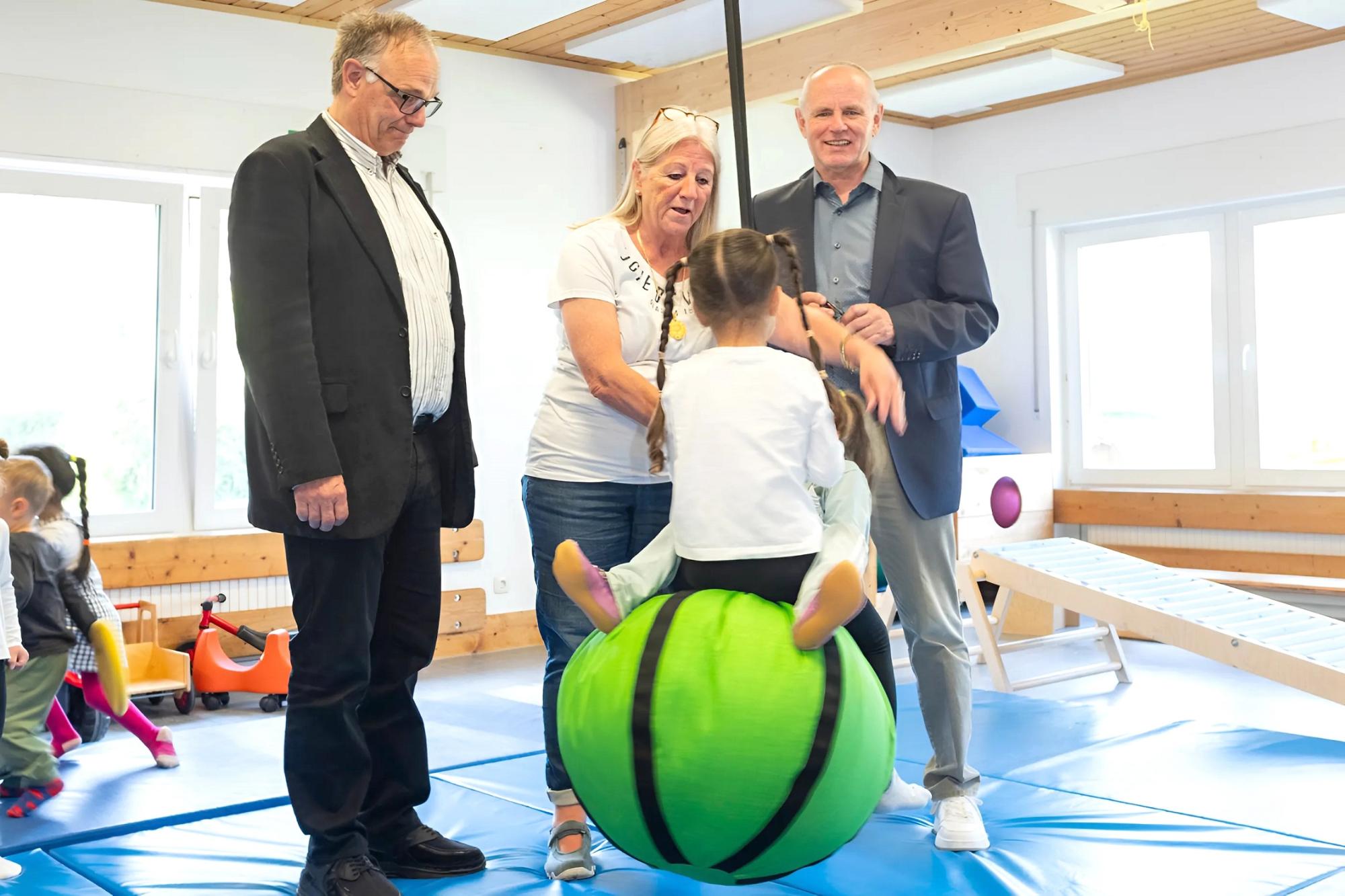 Kinder fliegen durch die Luft angeschubst von Kita-Leiterin Beate Hausmann-Solh. Ralf Claßen (r.) und Dr. Michael Stöhr freuen sich, dass mit der Spende der Mechernich-Stiftung der Schaukelball angeschafft wurde.