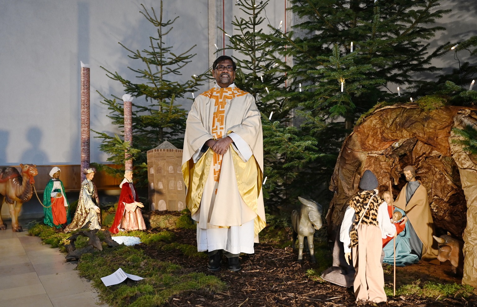 Pfarrer Stephen Rayappan Packiam inmitten der diesjährigen Dreikönigskrippendarstellung in der Pfarrkirche St. Johannes Baptist. Links bei der heranziehenden Karwane sind Malakowturm und die Hüttenschornsteine „Langer Emil“ und „Kurzer Karl“ vom früheren Bergwerksgelände „Op Spandau“ zu erkennen, die Küster Marco Sistig und Gebäudemanager Mario Fuß diese Weihnachten erstmals hinzugefügt haben. (c) Foto: Sabine Roggendorf/pp/Agentur ProfiPress